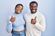 © Krakenimages.com - Young african american couple standing over blue background doing happy thumbs up gesture with hand. approving expression looking at the camera showing success.
