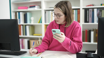 Wall Mural - Young blonde woman student using smartphone writing on notebook at library university