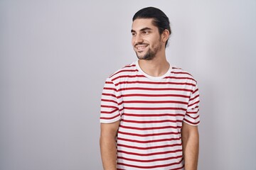 Wall Mural - Hispanic man with long hair standing over isolated background looking away to side with smile on face, natural expression. laughing confident.