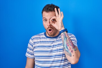 Canvas Print - Young hispanic man standing over blue background doing ok gesture shocked with surprised face, eye looking through fingers. unbelieving expression.