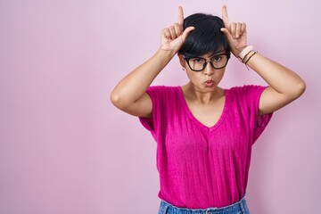 Canvas Print - Young asian woman with short hair standing over pink background doing funny gesture with finger over head as bull horns