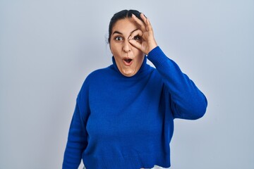 Sticker - Young hispanic woman standing over isolated background doing ok gesture shocked with surprised face, eye looking through fingers. unbelieving expression.