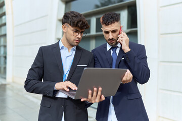 Wall Mural - Two hispanic men business workers using laptop talking on smartphone at street