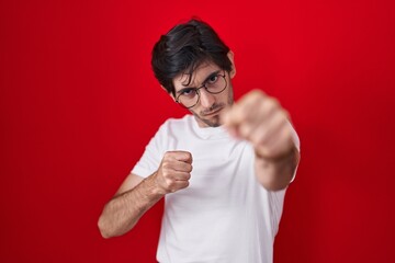 Poster - Young hispanic man standing over red background punching fist to fight, aggressive and angry attack, threat and violence