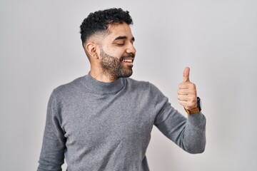 Poster - Hispanic man with beard standing over white background looking proud, smiling doing thumbs up gesture to the side