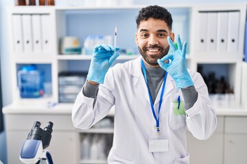 Poster - Hispanic man with beard working at scientist laboratory holding syringe doing ok sign with fingers, smiling friendly gesturing excellent symbol