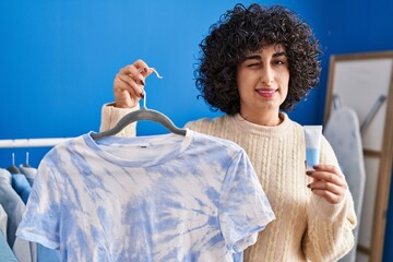 Poster - Young brunette woman with curly hair dyeing tye die t shirt winking looking at the camera with sexy expression, cheerful and happy face.