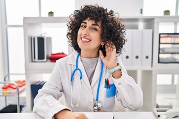 Sticker - Young brunette woman with curly hair wearing doctor uniform and stethoscope smiling with hand over ear listening and hearing to rumor or gossip. deafness concept.