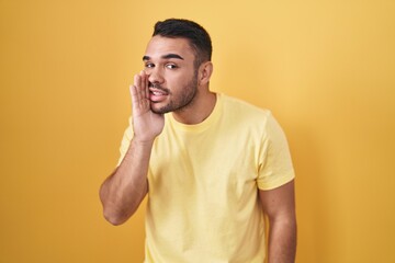 Poster - Young hispanic man standing over yellow background hand on mouth telling secret rumor, whispering malicious talk conversation