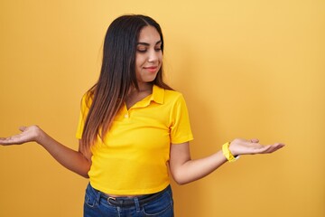 Wall Mural - Young arab woman standing over yellow background smiling showing both hands open palms, presenting and advertising comparison and balance