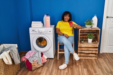 Wall Mural - Young beautiful latin woman using smartphone waiting for washing machine at laundry room