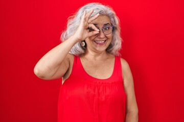 Poster - Middle age woman with grey hair standing over red background doing ok gesture with hand smiling, eye looking through fingers with happy face.