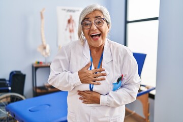 Canvas Print - Middle age woman with grey hair working at pain recovery clinic smiling and laughing hard out loud because funny crazy joke with hands on body.