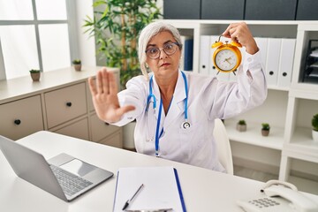 Poster - Middle age woman with grey hair wearing doctor uniform holding alarm clock with open hand doing stop sign with serious and confident expression, defense gesture