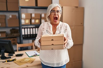 Poster - Middle age woman with grey hair working at small business ecommerce angry and mad screaming frustrated and furious, shouting with anger. rage and aggressive concept.