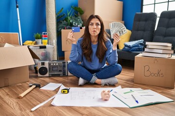 Sticker - Young brunette woman sitting on the floor at new home holding savings making fish face with mouth and squinting eyes, crazy and comical.