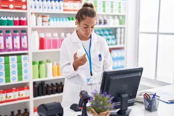 Young beautiful hispanic woman pharmacist using computer holding pills bottle at pharmacy