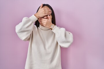 Wall Mural - Young south asian woman standing over pink background covering eyes and mouth with hands, surprised and shocked. hiding emotion