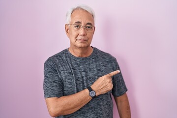 Sticker - Middle age man with grey hair standing over pink background pointing with hand finger to the side showing advertisement, serious and calm face