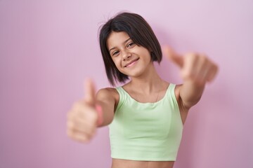 Wall Mural - Young girl standing over pink background approving doing positive gesture with hand, thumbs up smiling and happy for success. winner gesture.