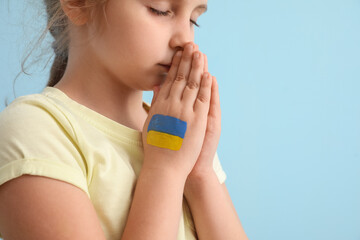 Little girl with drawn flag of Ukraine praying on blue background, closeup