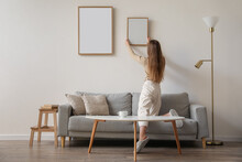 Young Woman Hanging Blank Frame On Light Wall At Home, Back View