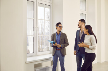 Male real estate agent showing new apartment to young couple. Happy customers getting tour through apartment ready to become homeowners. Happy clients and broker talking together looking at big window