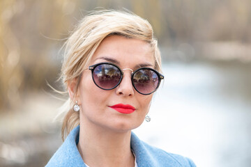 Street portrait of a stylish woman 40-45 years old in sunglasses on a blurry background of nature and water, close-up.