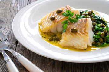 Fish dish - fried cod with spinach and capers in saffron sauce on wooden table
