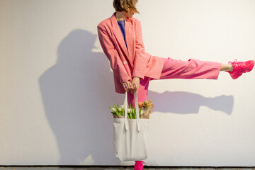 Wall Mural - Lifestyle portrait of stylish woman in pink suit with eco handbag full of flowers on white background. Canvas bag with blank space. Concept of style and spring time