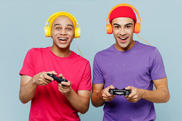 Wall Mural - Young exited cool couple two friends men wear casual clothes headphones together hold in hand play pc game with joystick console isolated on pastel plain light blue cyan background studio portrait.