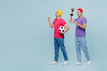 Full body young fan couple two friend men wear casual clothes together cheer up support football sport team holding soccer ball cup watch tv live stream isolated on plain pastel light blue background.