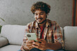 © ViDi Studio - Young Indian man wearing casual clothes use mobile cell phone listen music in earphones sits on grey sofa couch stay at home hotel flat rest relax spend free spare time in living room. Lounge concept.