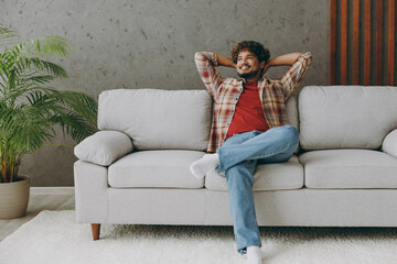 Wall Mural - Young fun Indian man wearing casual clothes hold hands behind neck looking aside sits on grey sofa couch stay at home hotel flat rest relax spend free spare time in living room indoor. Lounge concept.