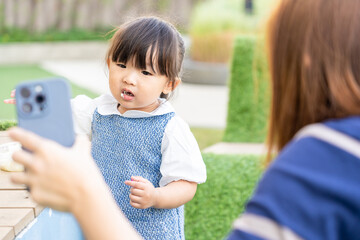 cute little girl with bangs hair eating snack and stained while playing outdoor in public park, kid take some food into mouth and vdo call with her father via mobile phone talk about holiday vacation