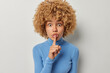 © WHstudio Leushin N - Surprised mysterious young woman with curly hair presses index finger to lips makes silence gesture dressed in casual blue turtleneck isolated over white background has secret. Be quiet sign.