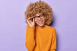 © WHstudio Leushin N - Portrait of happy positive European woman with curly hair laughs joyfully expresses happiness keeps hand on rim of spectacles dressed in casual orange jumper isolated over purple background.
