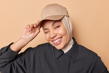 Wall Mural - Portrait of happy positive young woman winks eye smiles toothily feels very glad keeps hand on cap dressed in stylish clothes expresses nice authentic emotions isolated over beige background.