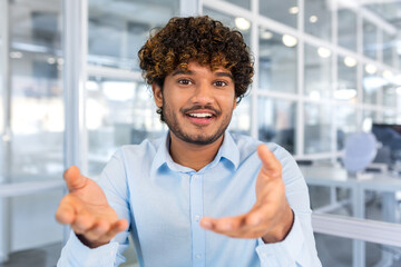 Video call online conference meeting, young successful hispanic man talking to colleagues remotely sitting in modern office, man looking at camera and smiling talking to colleagues in blue shirt.