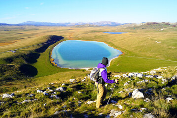 A back-turned hiker with hiking sticks stands in front of a beautiful blue lake in the middle of a grassy plain. Active holidays in Montenegro.