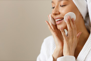 Wall Mural - Young woman in white robe and towel on her head wipes the skin of her face with cotton pads. Cleansing and healthy body care after shower. Close-up of pretty girl on isolated background, copy space.