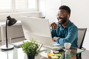 Handsome black male entrepreneur sitting at desk in office, looking at computer monitor and smiling, having video call