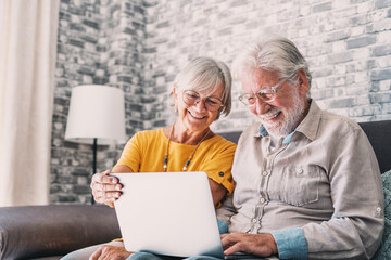 Pretty elderly 70s grey-haired couple resting on couch in living room hold on lap laptop watching movie smiling enjoy free time, older generation and modern wireless technology advanced users concept.