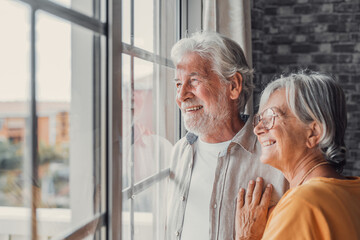 happy bonding loving middle aged senior retired couple standing near window, looking in distance, re