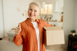 Stylish elderly female with short haircut posing with carton box showing thumb up with smiling facial expression, giving excellent mark for delivery service and quality of goods. Customer's feedback