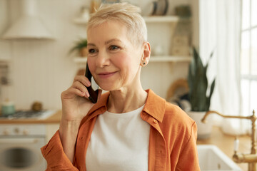 Portrait of adorable stylish retired lady in orange shirt talking on phone with her friend in kitchen, sharing gossips and rumors, last news, enjoying nice conversation with close mate