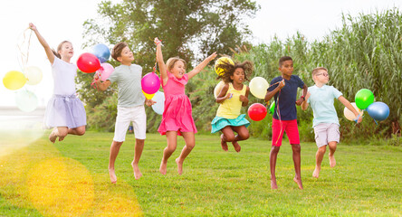 Wall Mural - Young joyful boys and girls jumping on lawn with balloons in hands.