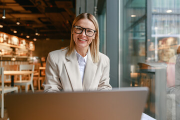 Wall Mural - Smiling businesswoman working on laptop while sitting in cafe near window and looking at camera