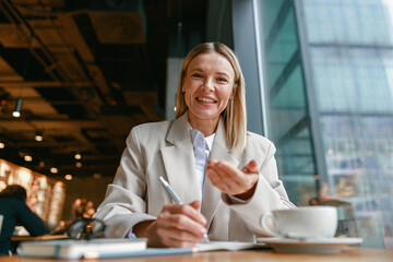 Wall Mural - Smiling businesswoman making notes and talks to camera while working in cafe