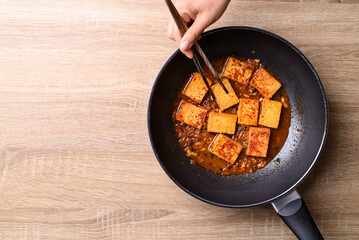 Wall Mural - Spicy braised tofu (Dubu Jorim) in cooking pan with hand and chopsticks, Korean side dish, Table top view